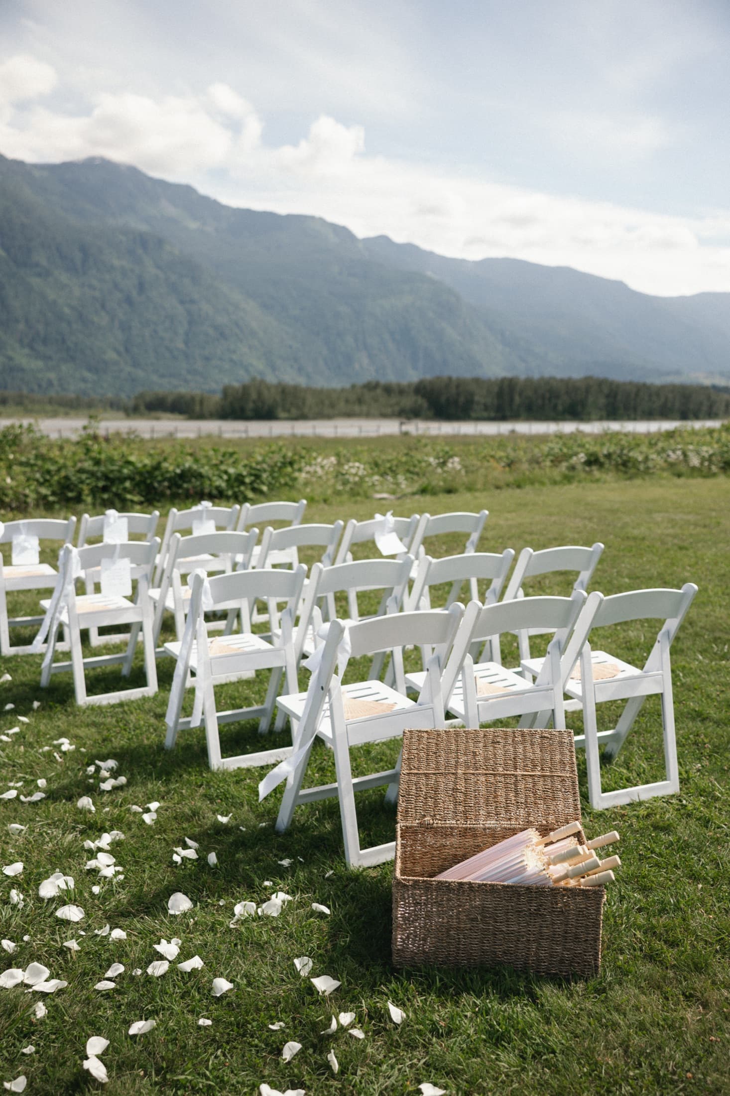 Rows of white folding chairs on grass with scattered petals and a woven basket of paper parasols, mountains and river beyond.