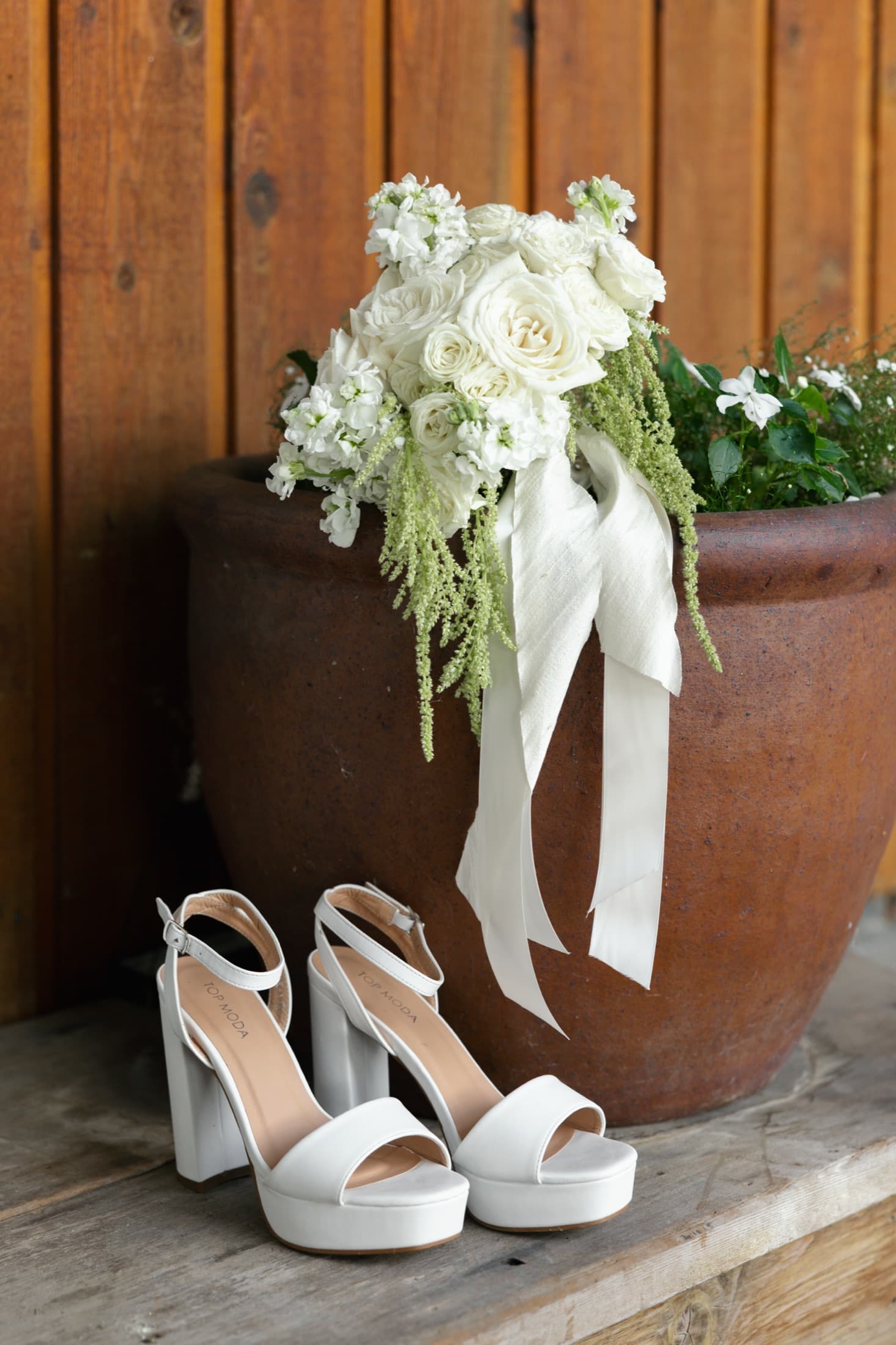 A white bouquet of roses and stock with trailing amaranthus and silk ribbon, resting on a terracotta planter above a pair of white heels.