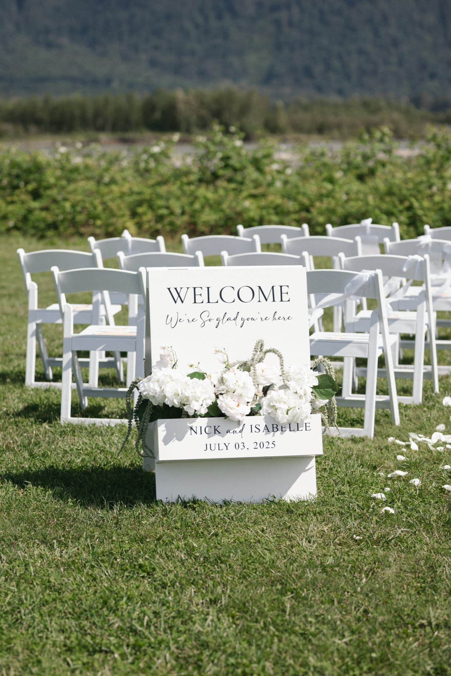 A white welcome sign on a plinth dressed with hydrangeas, standing at the head of an aisle of white chairs.