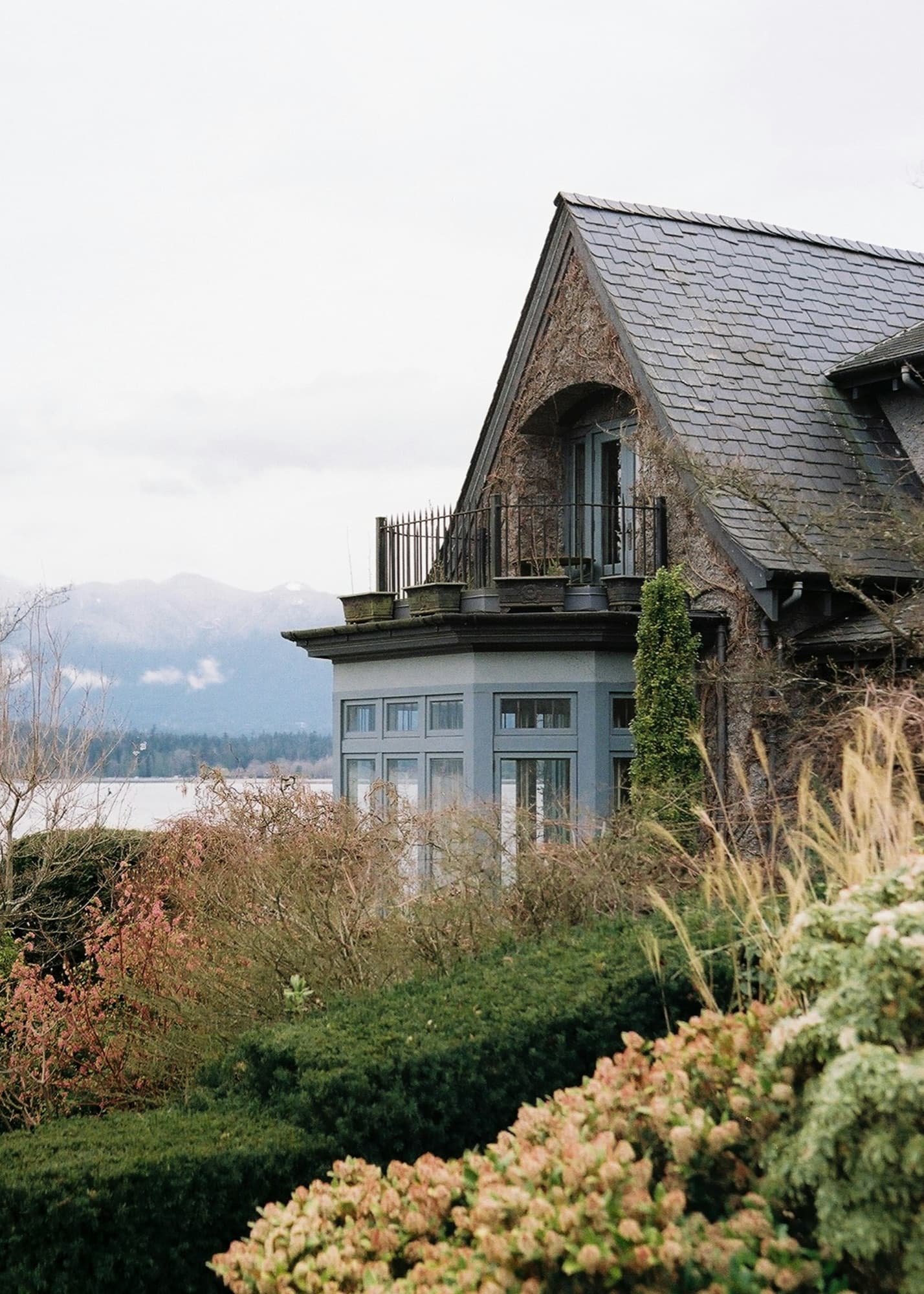 A heritage house with a steep slate roof and ivy-covered gable above a planted garden, water and mountains behind.