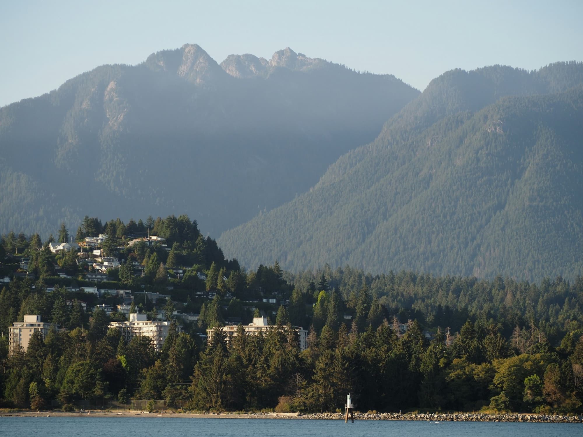Forested mountains rising above a wooded shoreline of houses, seen across open water in soft afternoon haze.