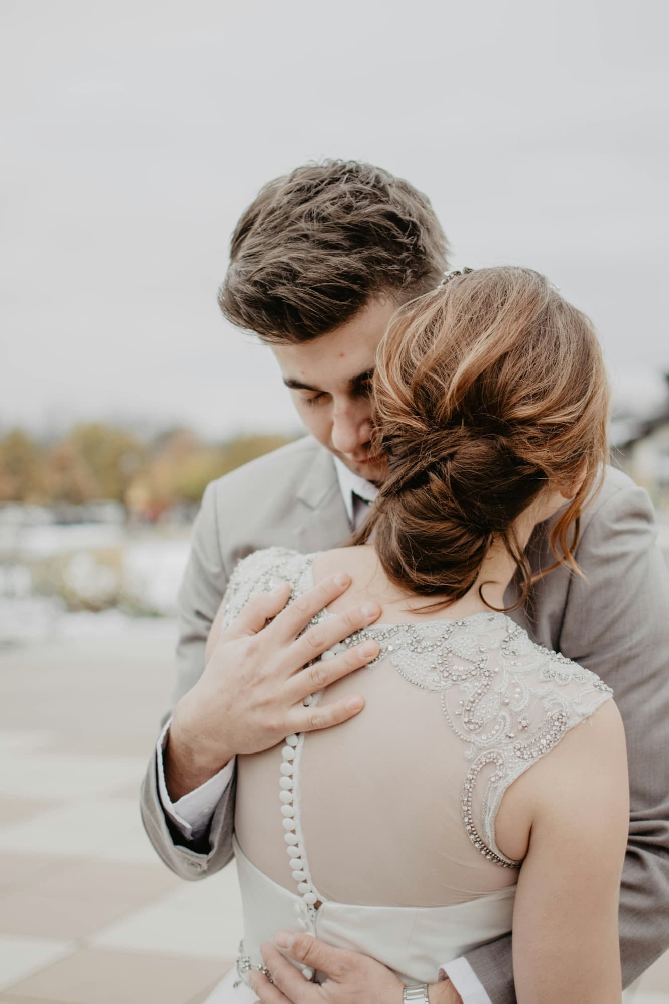 A couple holding each other quietly outdoors, his hand on her shoulder, the beaded back of her dress in focus.