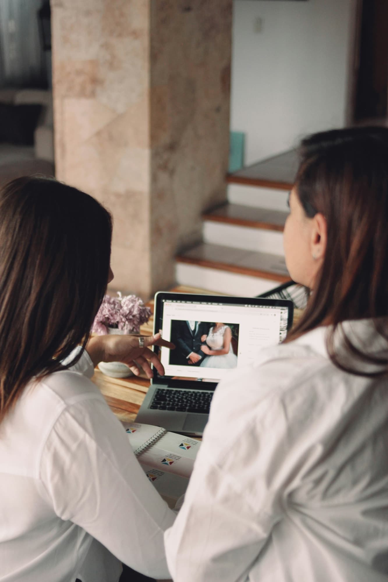 Two people sitting together at a laptop looking through wedding photographs, a notebook and flowers on the table beside them.