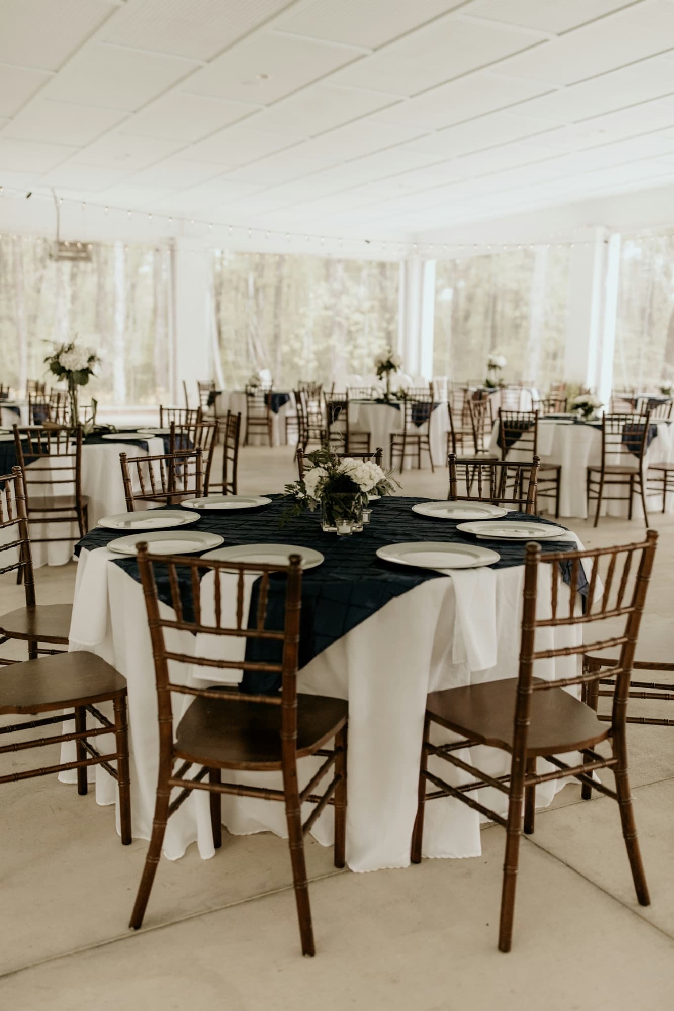 Round tables under a white pavilion, dressed with navy overlays, timber chiavari chairs and small white arrangements, woodland beyond.